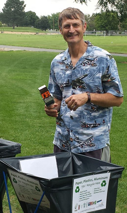 Photo of a man in a Hawaiian shirt stands over a recycling bin in park of green grass.