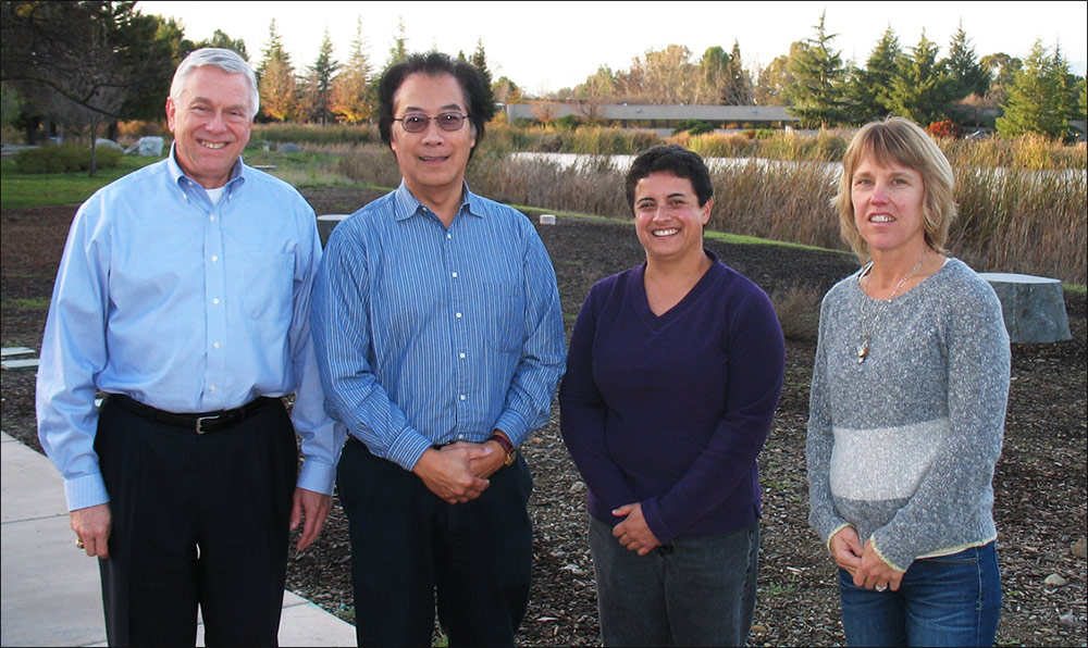 Photo of two smiling men and two smiling women stand in a row on a sidewalk in front of a garden space.