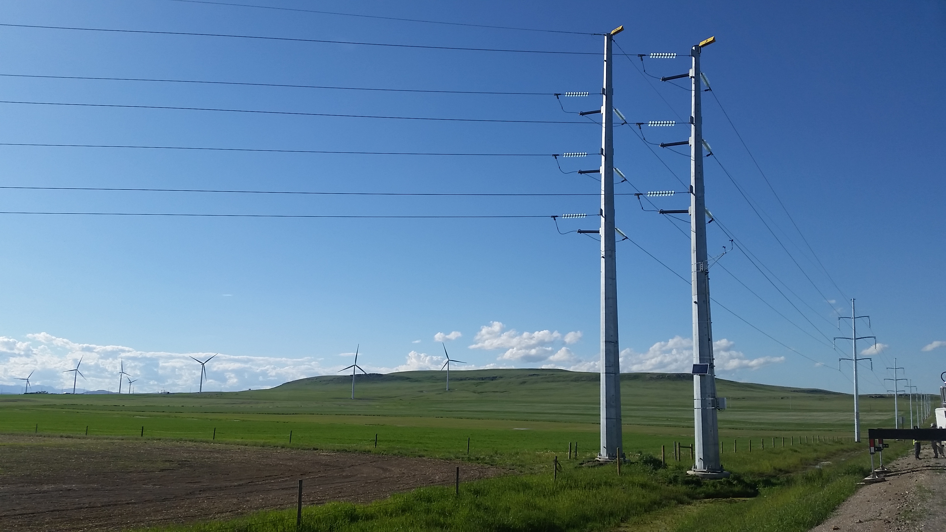 To increase the amount of electricity delivered from wind farms, researchers from the Energy Department and Idaho National Laboratory are investigating the effects of weather on transmission lines using weather stations and loggers, which are visible near the base of the utility pole on the right. <em>Photo from Idaho National Laboratory</em> Photo of two utility poles on open landscape with several wind turbines in the background