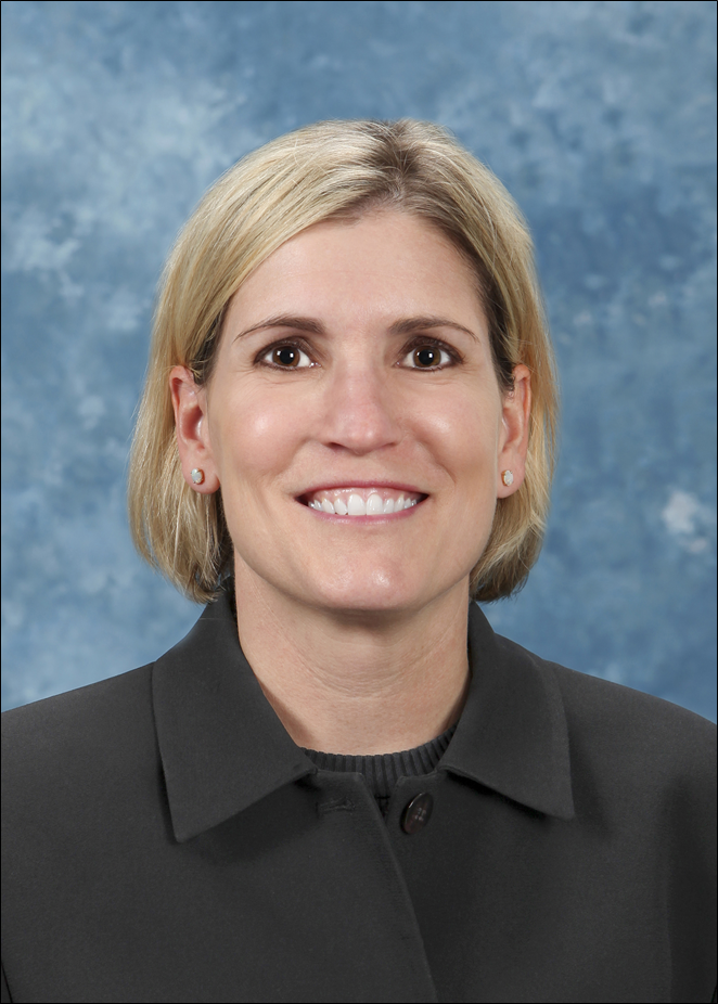 A headshot of a smiling blonde-haired woman with brown eyes wearing a professional gray suit. The background is light blue.