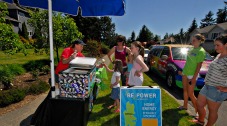 Photo of an outreach event: a woman at a cart with an umbrella, interacting with several people standing nearby, and a sign and car on the curb.