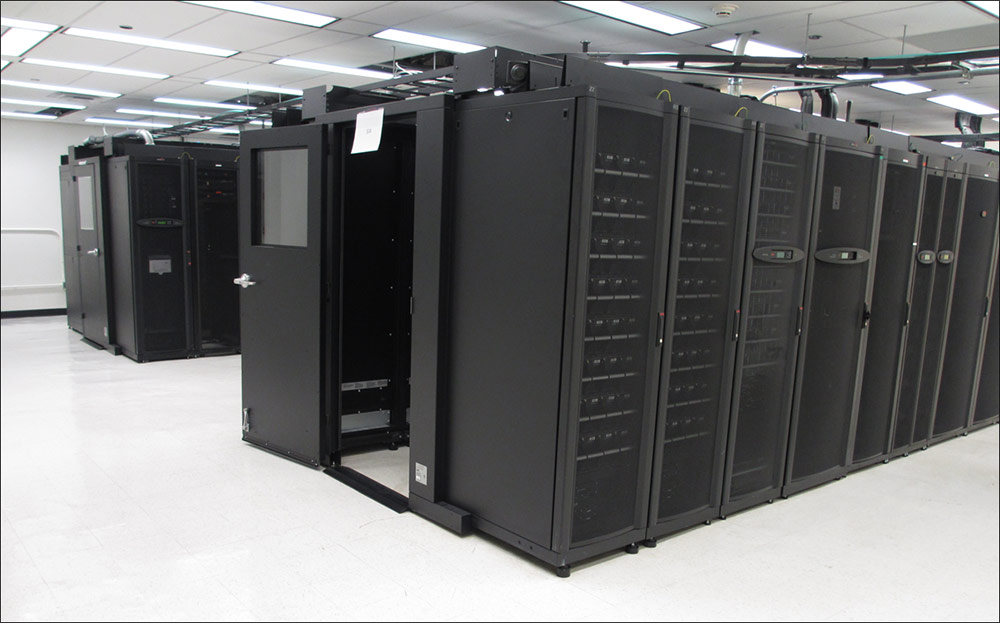 Photo of two long rows of black servers are situated in the middle of a data center that has white walls and a white suspended ceiling.