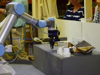The robot removes the sample from the clear transfer box, out of the two white sample containers, and transports it to the mock-up examination instrument. | Photo courtesy of Idaho National Lab. The robot removes the sample from the clear transfer box, out of the two white sample containers, and transports it to the mock-up examination instrument. | Photo courtesy of Idaho National Lab.