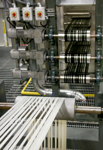 Pictured in the foreground, the white precursor (polyacrylonitrile, or PAN) fiber is being fed through the first oxidation oven. The black fiber behind that has passed through oxidation zones one and two and is passing through zone 3. At this point, it has turned black; during the oxidation process, the PAN precursor fiber gradually turns from white to yellow, auburn, brown, then black. Once fully oxidized, the fiber is ready to run through the higher-temperature furnaces, which convert the oxidized PAN fiber to carbon fiber. | Photo courtesy of Oak Ridge National Laboratory. Pictured in the foreground, the white precursor (polyacrylonitrile, or PAN) fiber is being fed through the first oxidation oven. The black fiber behind that has passed through oxidation zones one and two and is passing through zone 3. At this point, it has turned black; during the oxidation process, the PAN precursor fiber gradually turns from white to yellow, auburn, brown, then black. Once fully oxidized, the fiber is ready to run through the higher-temperature furnaces, which convert the oxidized PAN fiber to carbon fiber. | Photo courtesy of Oak Ridge National Laboratory.