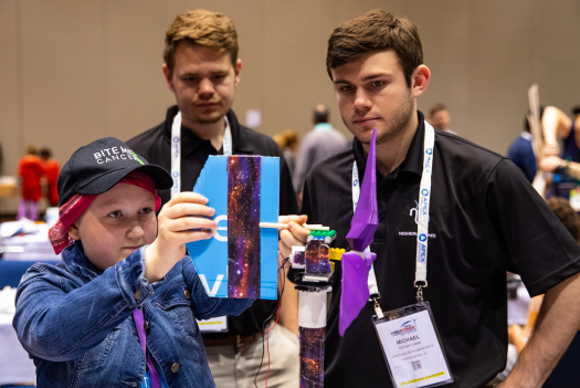 Two college students look on as a child shows off a wind turbine design.