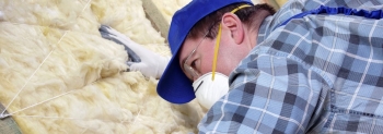 Photo of a man working on insulation in a building, wearing a hat and a face mask.
