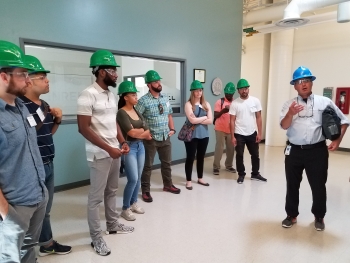 A grounp of young men and women with green hard hats on listen as a man in a blue hard hat talks and guestures at something to his right.
