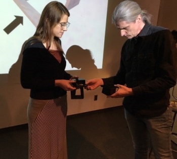 Photo of two people in a presentation room, with a screen behind them, examining objects for a project.