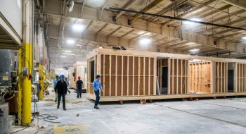 The inside of a large warehouse with a construction project going on and workers walking around it.