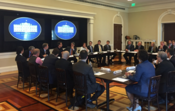 people seated at four tables facing the center in a White House room. 