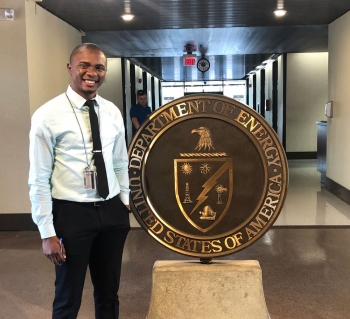 Johnson Okoduwa Imumbhon stands smiling next to a statue of the US Department of Energy seal