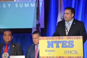 Honorable James E. Campos speaks at a podium labeled NTES. Kevin Frost and another man listen attentively.