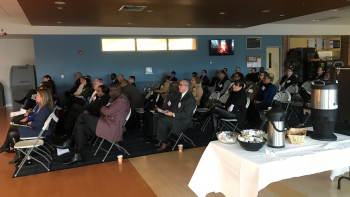 Men and women in business suits sit in rows of folding chairs. In the foreground is a coffee station.