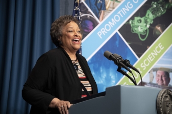 Kay Cole James is speaking at a podium. She is smiling. The DOE seal is partially visible on the podium. Visible in the background is the American flag.