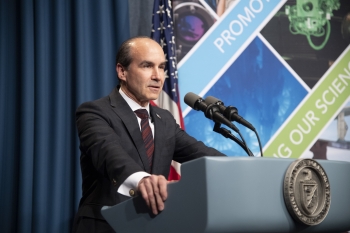 Under Secretary Menezes is speaking at a podium. Visible on the podium is the DOE seal. Visible in the background is the American flag.