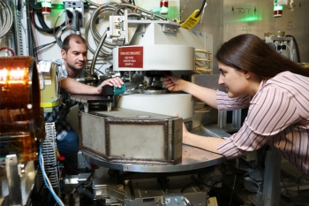 From left, ORNL staff Matthew Frost and Leah Broussard work at the Magnetism Reflectometer at the Spallation Neutron Source, used for a search for mirror neutrons. A woman and man are looking at and making adjustments to a piece of complex metal and plastic machinery with wires and tubes behind it.