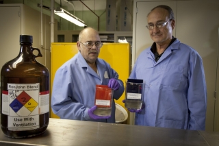 Photo of two men standing in laboratory wearing blue lab coats, holding beakers with a liquid in them. A jug of RonJohn Blend is on a table in the foreground.