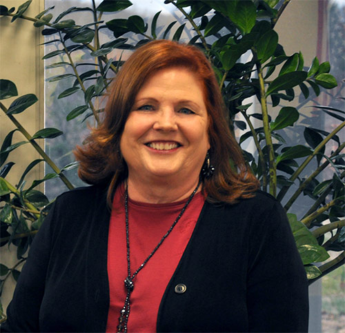 Photo of a smiling woman in a red shirt and navy blue sweater standing in front of an indoor plant.