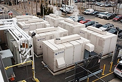 The Verizon Telecommunications Center in Garden City, New York, is powered by fuel cells. Photo of seven fuel cells with a series of pipes to the side in a parking lot setting. Steam is rising above some of the fuel cells.