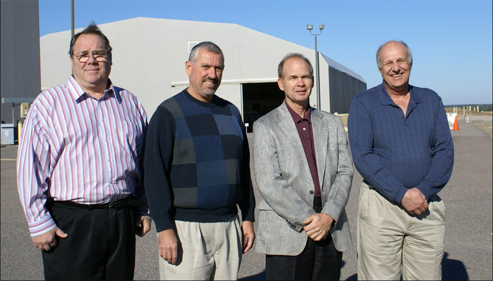 Photo of four men standing outside on a clear blue sky day. There are large gray hanger buildings in the background.
