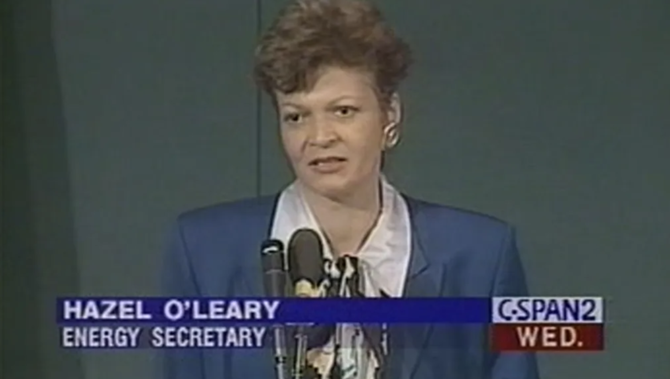 Video screengrab of original CSPAN footage as Secretary O'Leary announces the Openness Initiative. She is standing in front of a microphone where a blue suit jacket and white collared shirt.